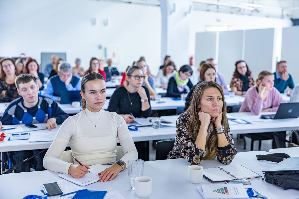 V popředí sedí dvě ženy, které si zapisují poznámky a sledují program.