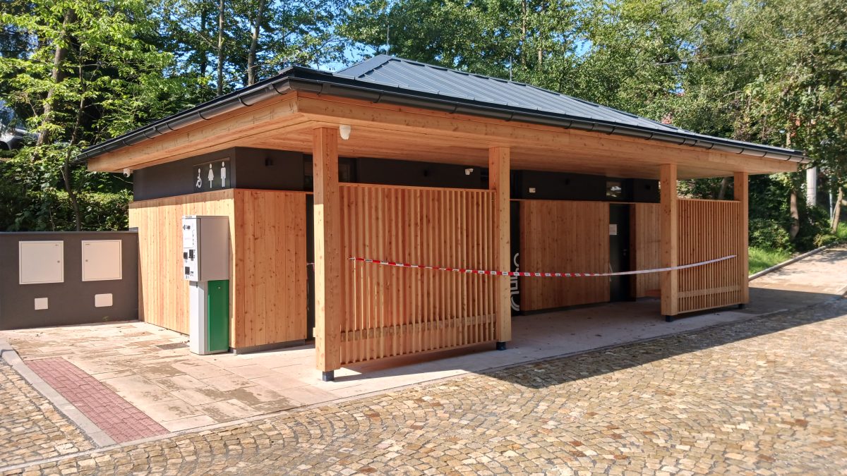 Moderní dřevěný objekt veřejných toalet s bezbariérovým přístupem, umístěný na dlážděné ploše u lesa.