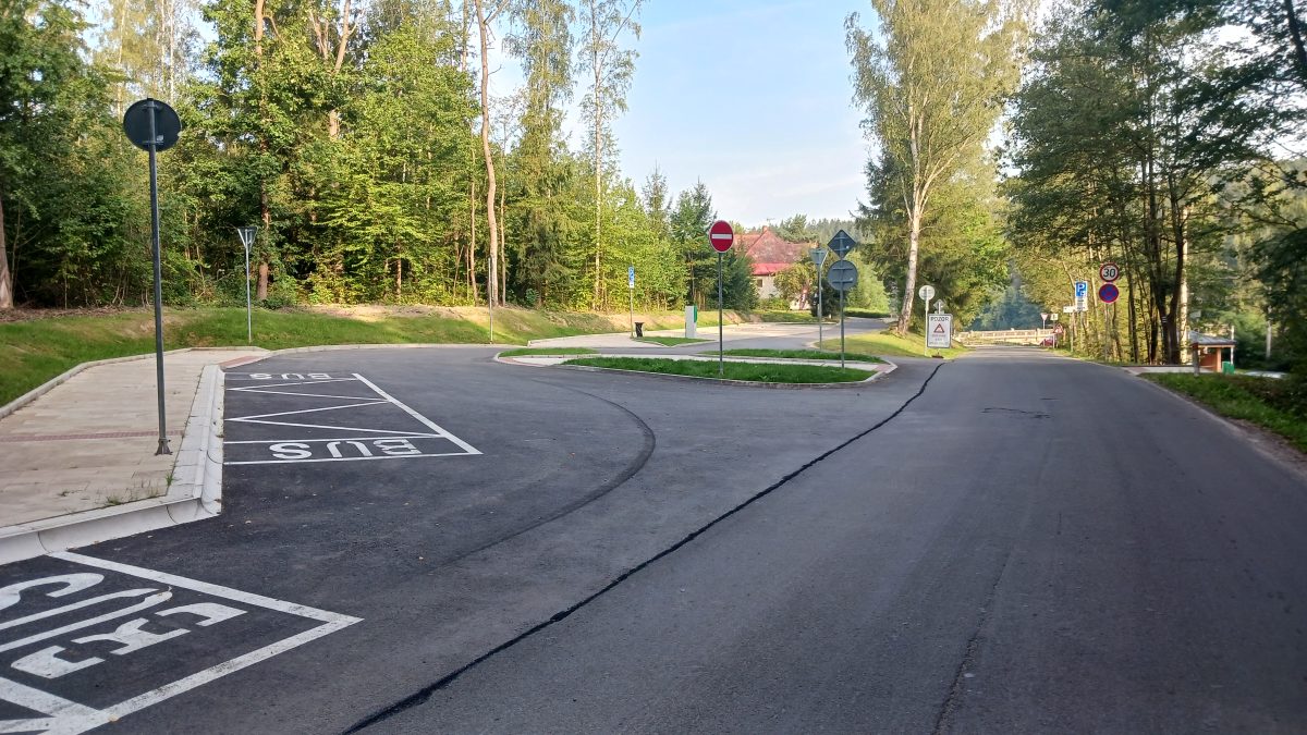 Asfaltová komunikace u lesa s vyznačenými parkovacími místy pro autobus, dopravními značkami a chodníkem po levé straně.