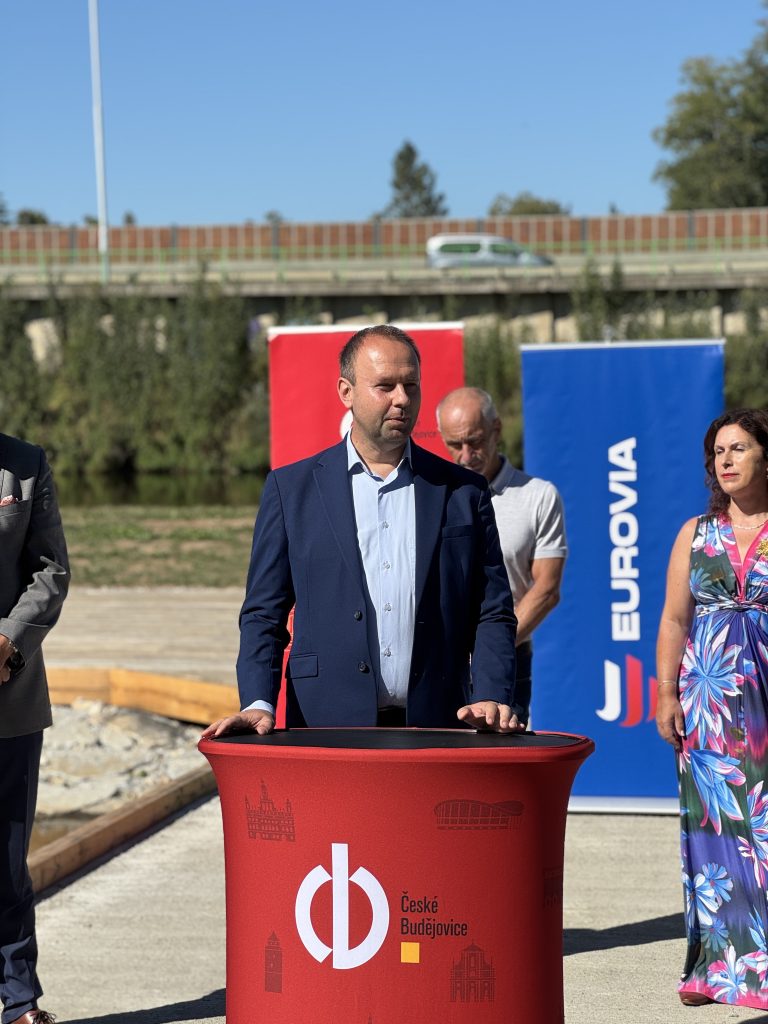 Petr Bouška mluvící u oficiálního pultu s logem Českých Budějovic, v pozadí hosté, voda a modré i červené bannery.