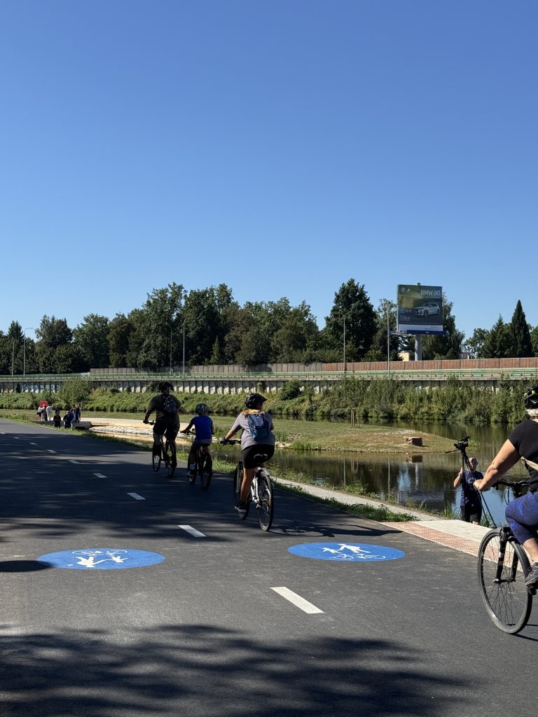 Cyklisté všech věkových kategorií na nové asfaltové cyklostezce u Vltavy, v pozadí chodci a Sluneční ostrovy.