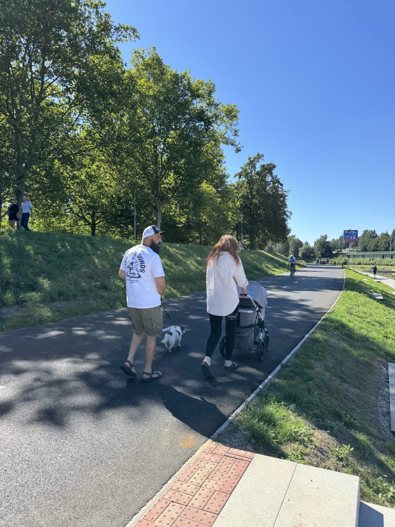 Mladá rodina se psem a kočárkem na nově vybudované stezce podél řeky, stín stromů a upravený terén kolem cesty.