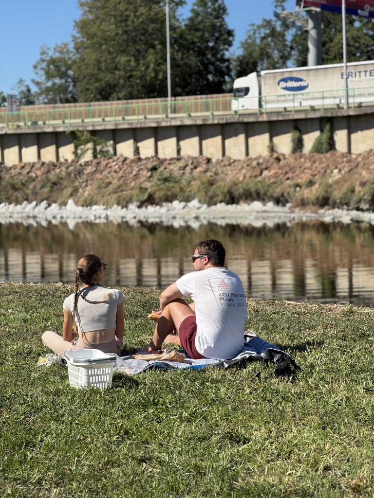 Muž a žena sedící na dece u řeky Vltavy během slunečného dne. Piknik na trávě, v pozadí nábřežní zeď, kamení a projíždějící kamion.