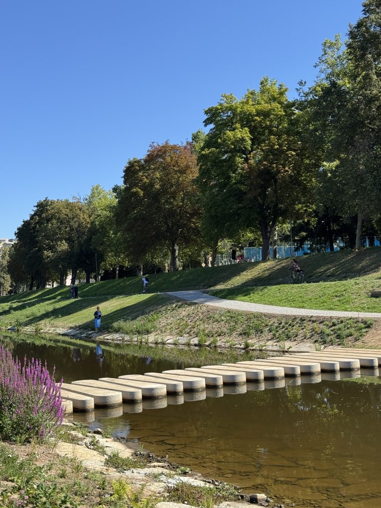 ohled na betonové šlapáky vedoucí přes řeku, obklopené zelení, stromy a upraveným břehem. Modrá obloha a lidé v dálce na cestě.