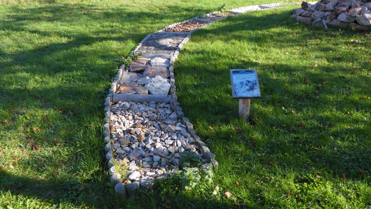 Úsek naučné stezky z různých druhů kameniva s informační tabulkou, vedoucí přes travnatý pozemek.