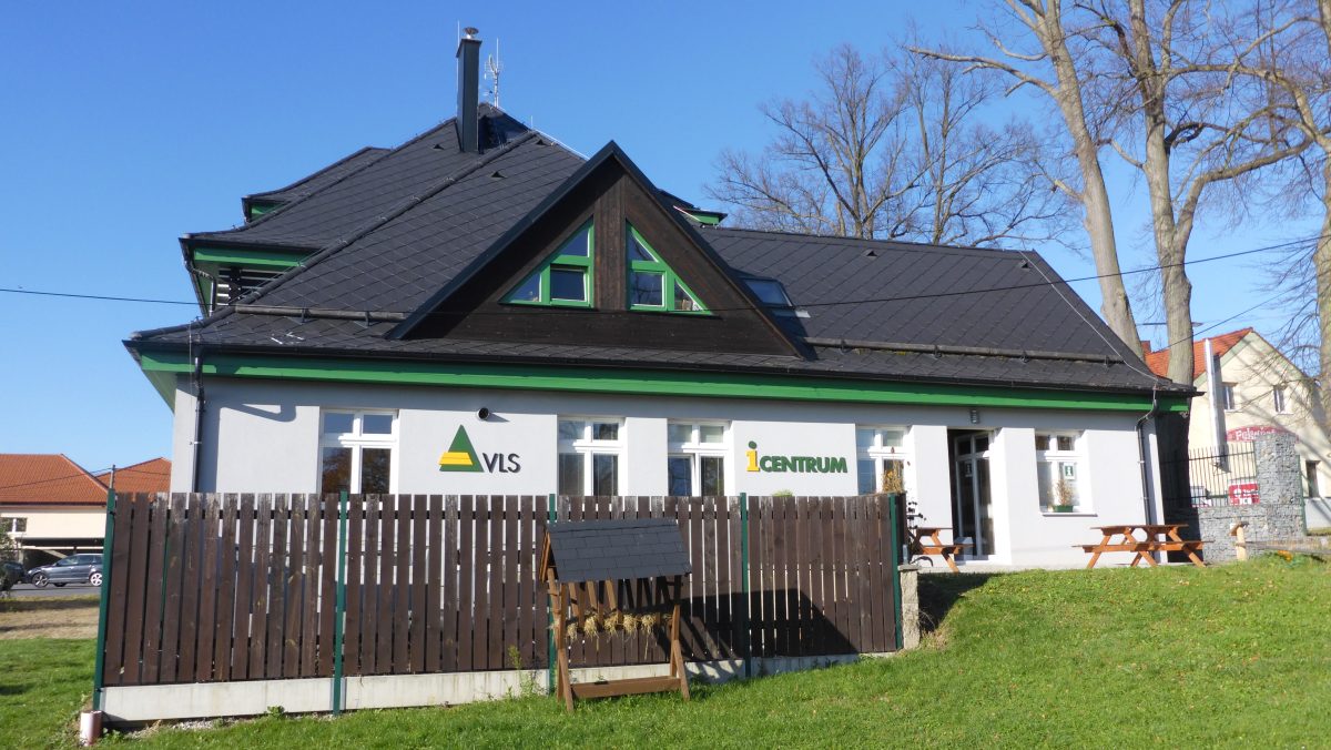 Budova turistického informačního centra VLS s šedou fasádou a černou střechou, před níž stojí dřevěný plot a lavičky.
