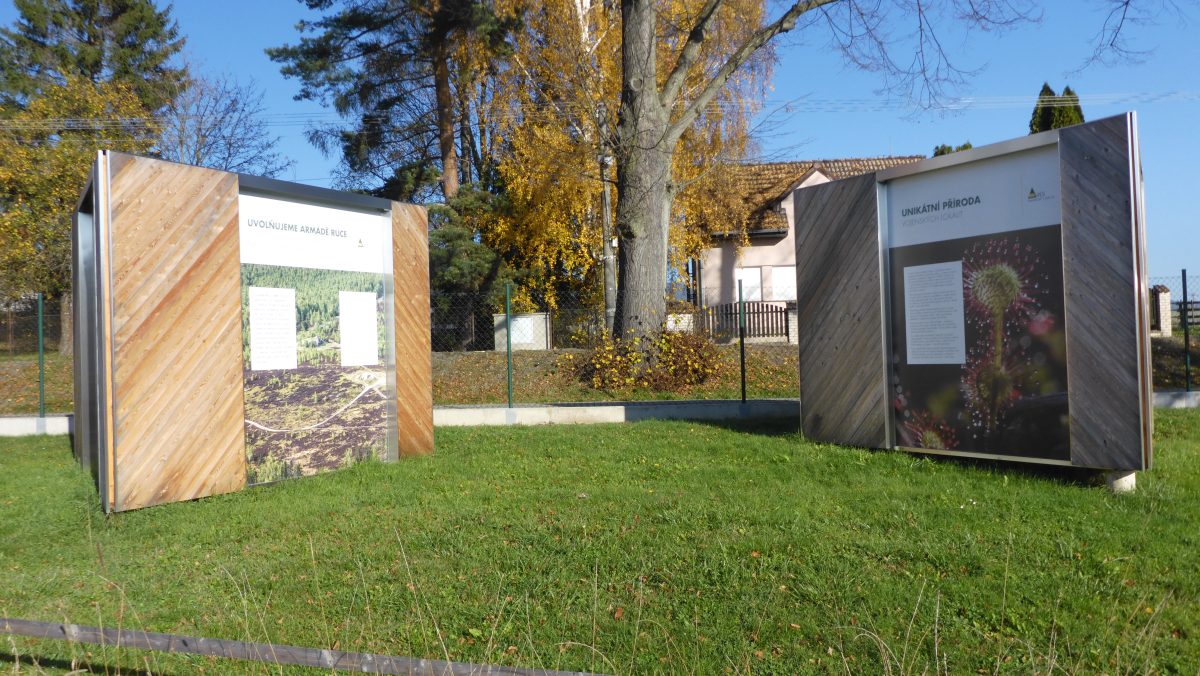 Dva venkovní informační panely s dřevěným rámem a fotografiemi, umístěné na travnaté ploše s podzimními stromy v pozadí.