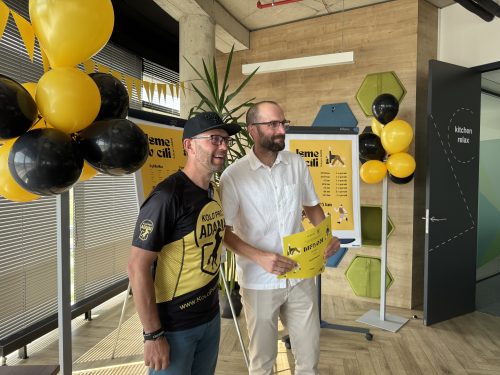 Účastník přebírá diplom, společně s hostem pózují u výzdoby z balónků a výsledkové tabule s nápisem „Jsme v cíli“.