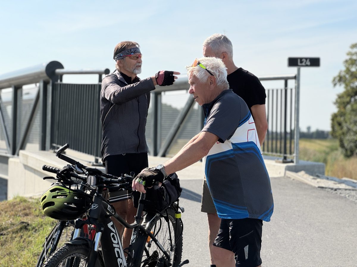 Dvojice starších cyklistů diskutuje u mostu, vedle nich stojí kola.