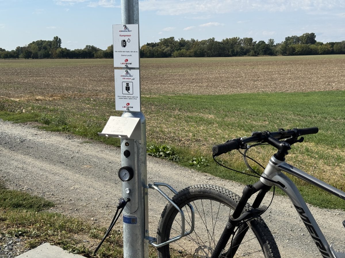 Cyklostojan s pumpou a USB nabíječkou na stezce, vedle odstavené kolo.