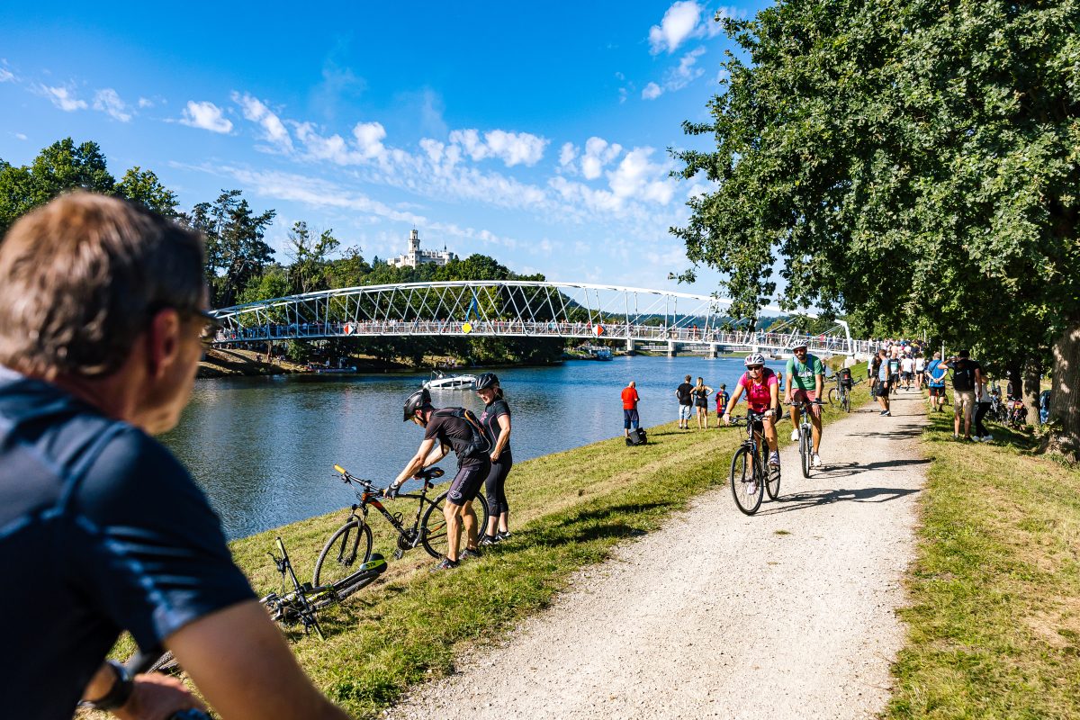 Cyklisté a návštěvníci na břehu Vltavy s pohledem na novou lávku v Hluboké