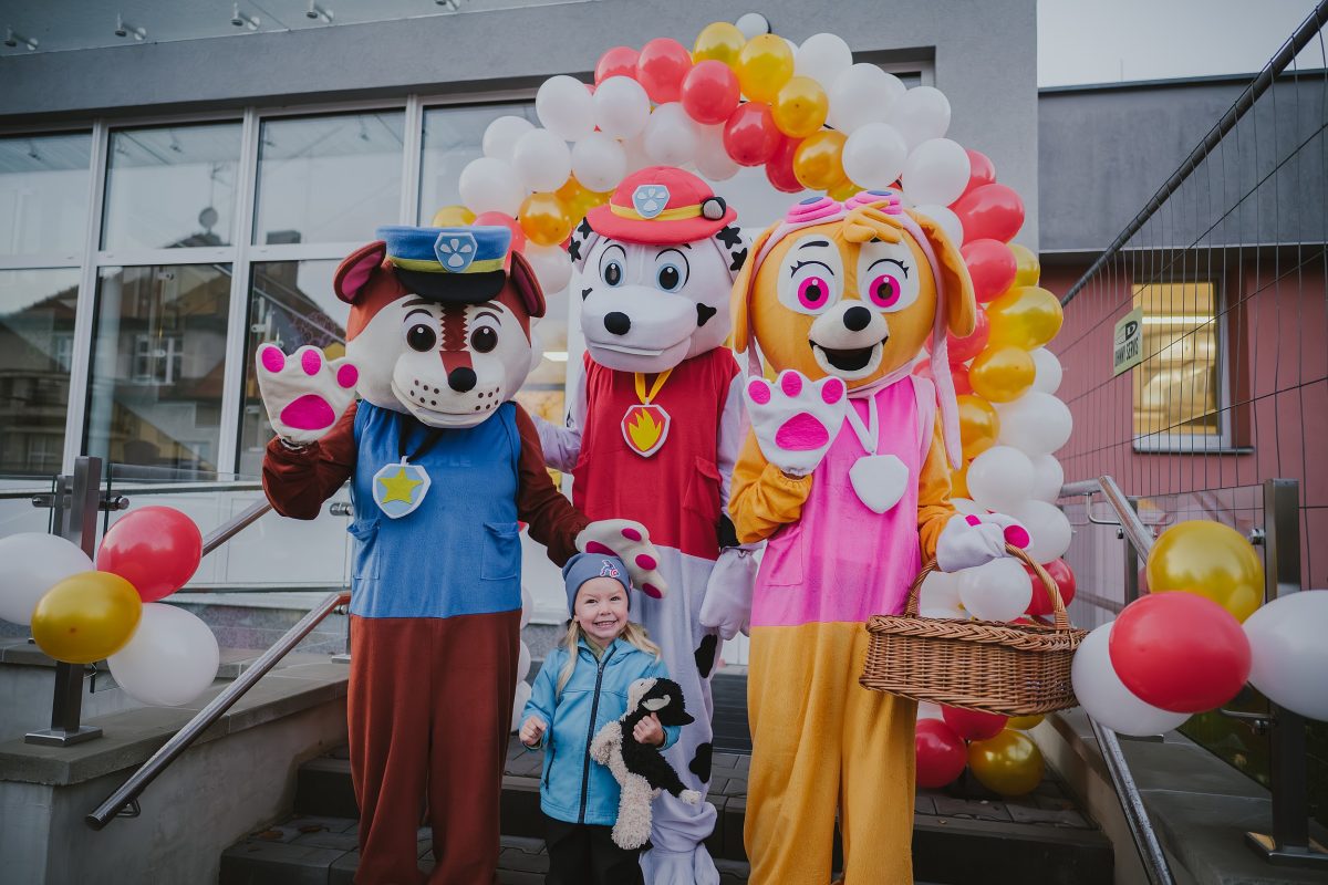 Děti pózují s maskoty před vstupem nové mateřské školy Zavadilka ozdobeným balónky.