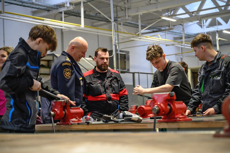 Studenti pracují v dílně u pracovního stolu se svěráky pod dohledem instruktorů.