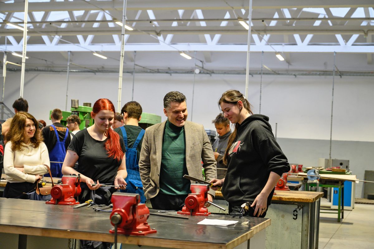 Studenti pracují v dílně u pracovního stolu se svěráky pod dohledem instruktorů.